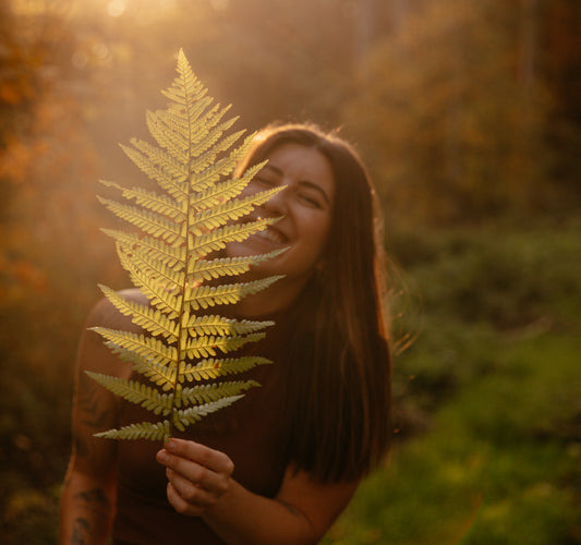 Angie hält im Wald einen Farn vor sich und lächelt. 